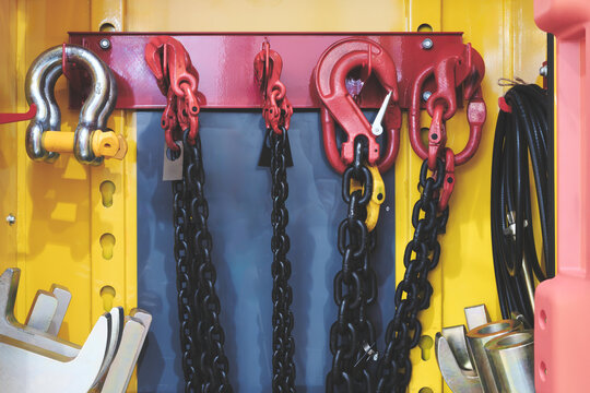 Yellow wall supports a red metal bar. Red chains hang from hooks on the bar. Black chains dangle below the red ones. Yellow chain hook stands out prominently. Black cables coil on the wall