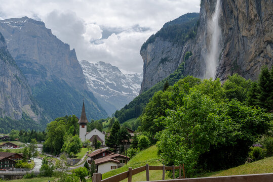 Summer landscape of Swiss alpine valley with church, traditional chalets, lush trees, and a tall waterfall beneath dramatic mountain cliffs.