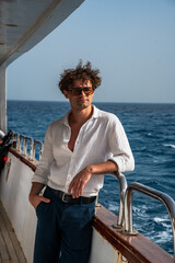 Fototapeta premium Epic tropical cruise traveler man luxury yacht vertical portrait. Cinematic full length view of confident wealthy male explorer on boat deck against blue ocean water and horizon sky. 