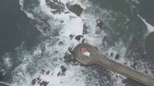 A drone view of Felgueiras Lighthouse perched on a stone pier surrounded by crashing waves and rocks in misty ocean waters in Porto, Portugal