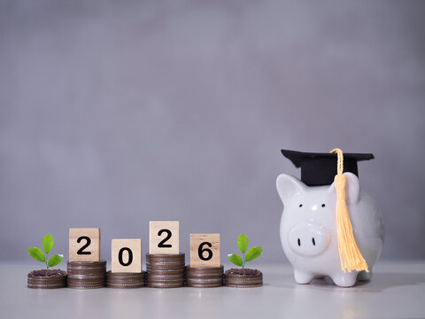 Piggy bank with graduation hat, Wooden block with number 2026 and plants growing on stack of coins. The concept of saving money for education, student loan, scholarship, tuition fees in year 2026.