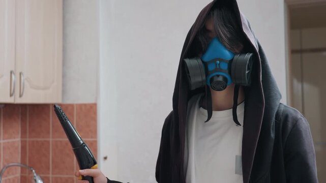 Masked cleaner in kitchen wearing respirator and hood hooded figure holds sprayer nozzle by sink and countertop during deep home disinfection, protective gloves and apron, tiled backsplash