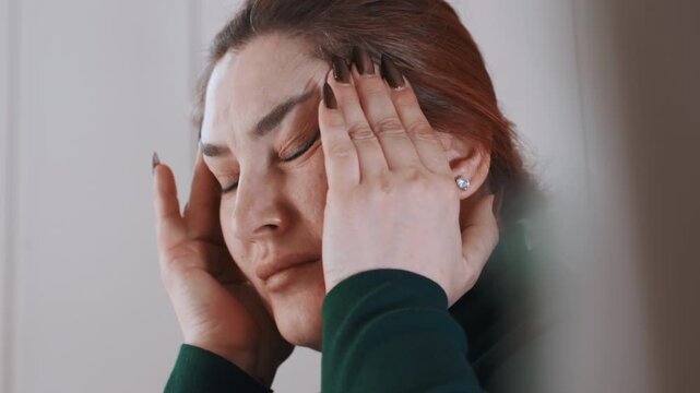 White woman clutching temples in pain at home, tight closeup showing flushed face and tense expression, fingers pressing temples, short nails, green sweater, intimate mood conveying migraine,
