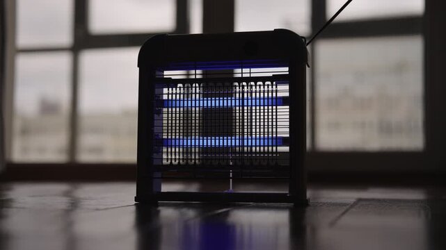 Electric insect killer lamp turned on, glowing with blue ultraviolet light to attract and kill bugs. The device switched off, leaving it in silhouette against window, close-up, slow motion.