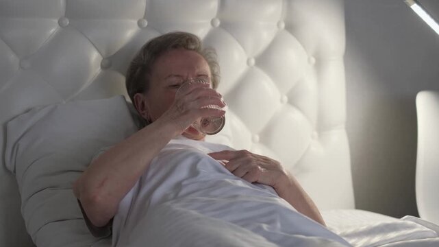 A sick elderly woman drinks water in bed at night