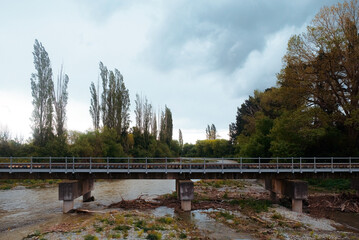 Fototapeta premium South Island, New Zealand: Scenic roadtrip view of concrete bridge crossing shallow gravelly riverbed below tall slender poplar trees under cloudy sky.