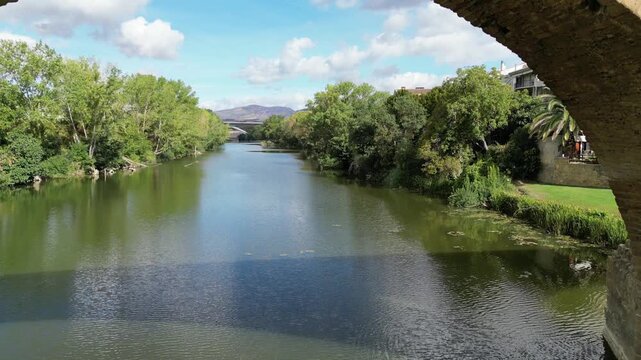 A drone view of the ancient Puente la Reina stone bridge crossing Arga River, with trees, buildings, and distant mountains under a bright sky in Spain