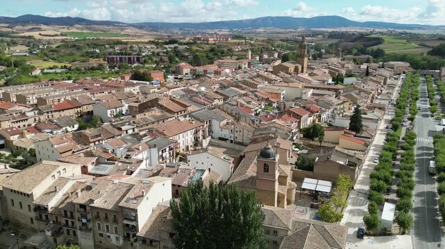 A drone view of Puente La Reina historic town with tiled rooftops, church towers, tree-lined streets, and distant mountains under a bright sky, Spain