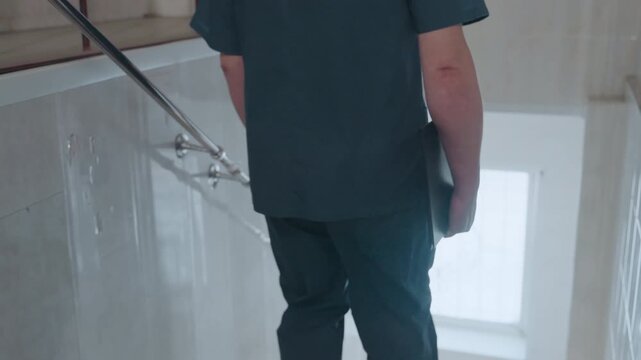 Paramedic walking down dim tiled stairwell, back view of nurse in scrubs carrying clipboard, slow descent toward landing of residential building, quiet mood and reflective posture after home