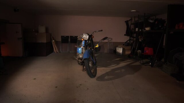 Vintage blue motorcycle sits under a lone spotlight in a dim garage, dust coating its chrome as shadows fall across shelves, boxes and concrete floor, evoking neglect and mystery