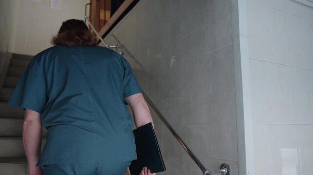 Medic ascends staircase. Paramedic in white uniform ascends tiled stairwell carrying tools. Health professional in white attire walks up tiled staircase with clipboard and stethoscope in hand