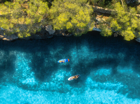 Aerial drone view of colorful kayaks in crystal clear turquoise sea near rocky coast and green trees. Summer outdoor adventure in Mallorca, Balearic Islands, Spain. Top down view of canoes. Vacation	