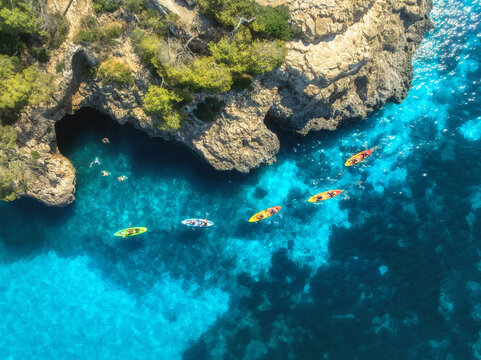 Aerial drone view of colorful kayaks in crystal clear turquoise sea near rocky coast and green trees. Summer outdoor adventure in Mallorca, Balearic Islands, Spain. Top down view of canoes. Vacation	
