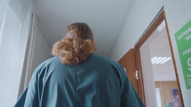 Clinic caregiver walking down bright hallway back view in blue scrubs with ponytail, passing patient room doors and informational posters, soft daylight through curtains, calm professional pace