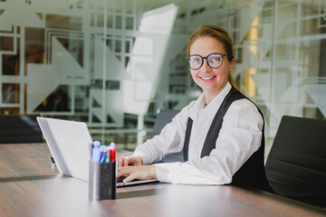 Fototapeta premium Professional woman working at her desk in a modern office setting while smiling and typing on a laptop during a bright afternoon