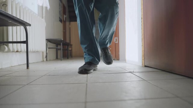 Black paramedic walking down hallway toward door, blue scrubs and black shoes on tile floor, lowangle shot showing bench and radiator, steady footsteps conveying urgency and focus while approaching