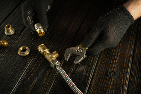 In a home setting, a person wears black gloves and uses a wrench on a plumbing part. Metal fittings and a flexible hose are also on the table. The scene shows work in progress