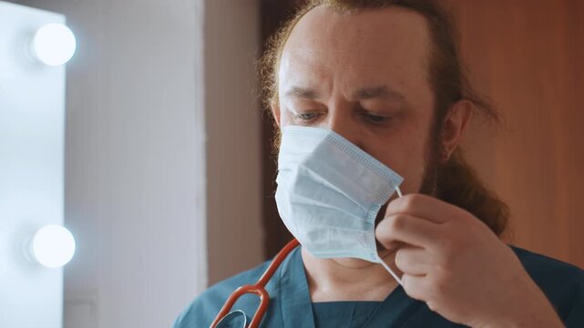 Mirror lit medic tightening mask, stethoscope on neck white male checks fit under vanity lights, secures ear loops, keeps steady gaze and calm professional demeanor, preparing for close contact care
