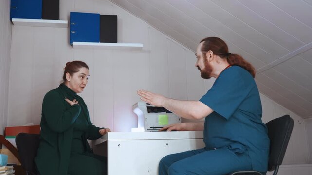 Paramedic home assessment at desk patient concerned seated near printer lamp clinician in blue scrubs reviewing records calm supportive interaction small attic office paperwork review vital check