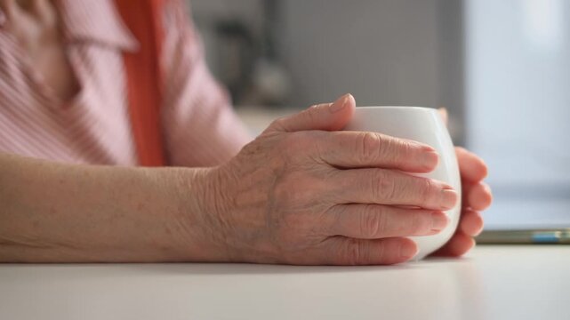 An elderly woman warms her hands on a cup of tea, close-up. Deterioration of blood flow in old age concept