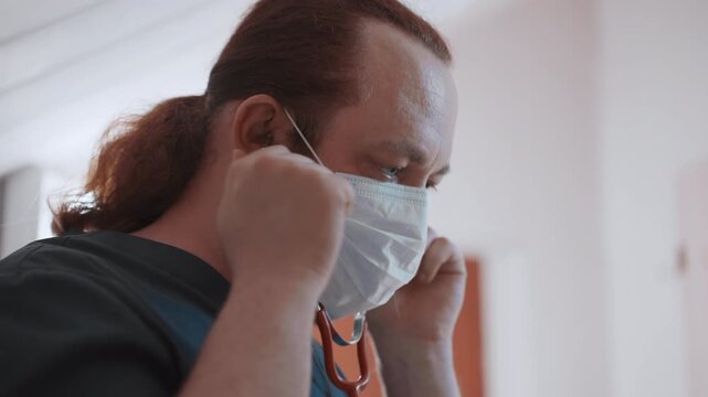 Masked clinician prepares stethoscope for exam, close profile of male medic wearing surgical mask and hair tied back, adjusts earpieces with focused concentration under soft clinical lighting, PPE