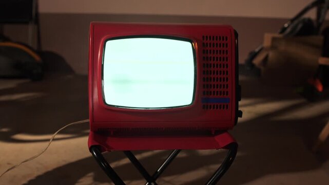 Retro red television with an empty, glowing static screen placed on a black stool in a dimly lit indoor setting, evoking nostalgia and outdated technology