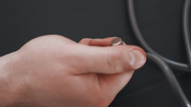 White paramedic hand holding stethoscope against dark background, closeup of metal chest piece and tubing during home visit, gentle adjustment and focused assessment, ambient soft lighting conveys