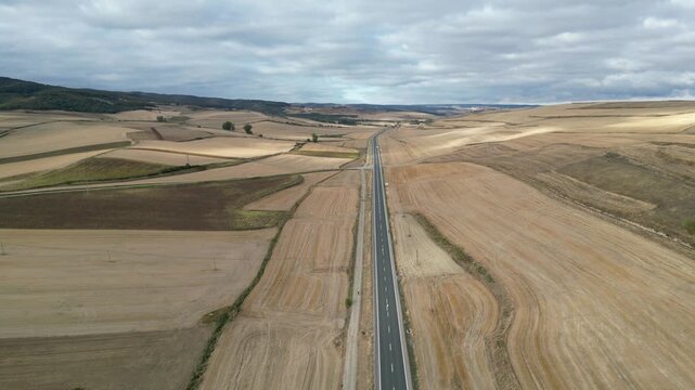A drone descending view of walkers following rural path beside a long straight road cutting through wide golden farmland at Camino de Santiago, Spain