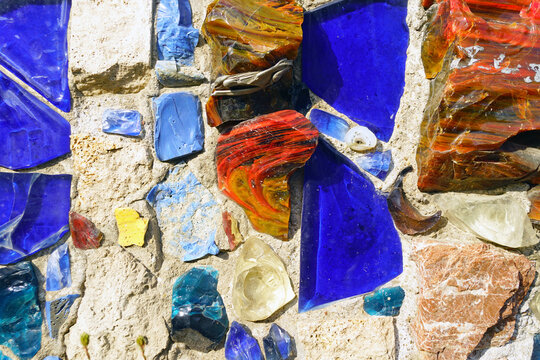 Cobalt blue glass shards and multicolored stones embedded in textured wall surface. Conveys creative placemaking, cultural revitalization, and artistic reuse through reclaimed-material design.