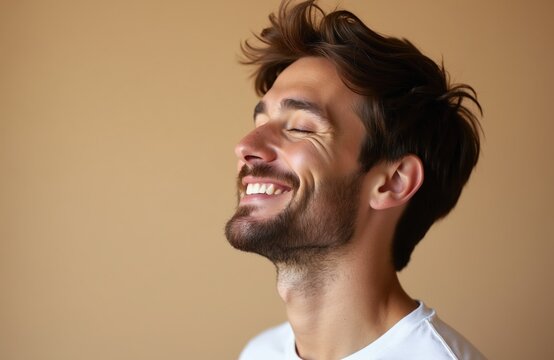 Man laughs with eyes closed. He smiles broadly showing teeth. Young man experiences joy. He feels happiness. Male person has beard. His head tilts back.