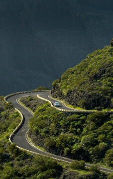 A white car drives past a sharp hairpin turn on a winding road through Masca in Tenerife, offering breathtaking panoramic views over the dramatic and lush green mountain landscape.