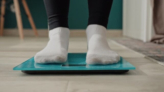 Morning home weighin on teal scale in hallway, person in white socks steps onto digital glass scale near rug and chair, bright interior light, focused tracking of weight progress and wellness habit