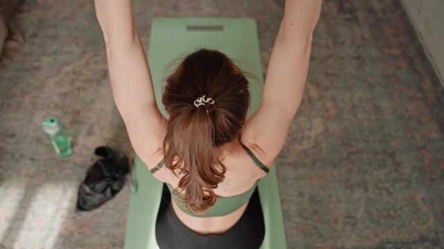 Caucasian woman house resistance workout seated on green mat pulling band overhead, workout gloves, water bottle and shoes nearby, overhead angle, controlled repetitions for shoulder and upper back