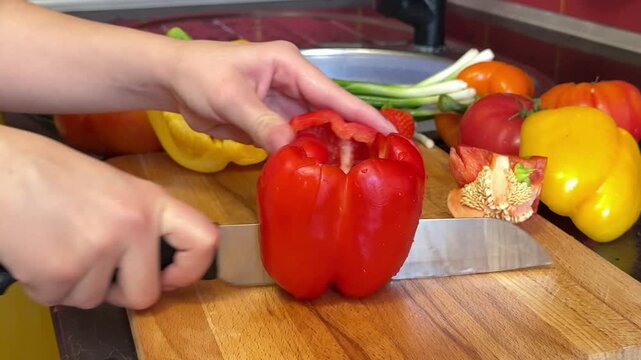 Female hands cutting juicy red bell pepper into halves with large knife and then slicing fresh vegetable into thin strips during homemade cooking preparation.