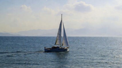 Fototapeta premium Sailboat moving across calm sea with distant mountains