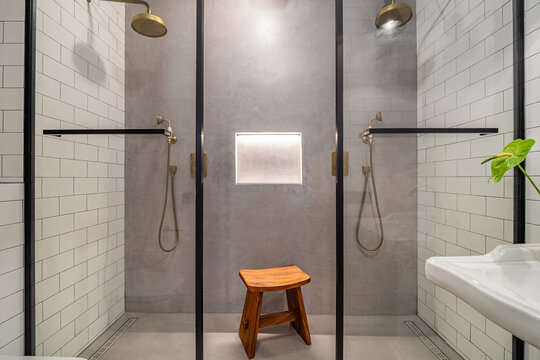 Modern bathroom with two glass shower stalls, a wooden stool, and a small green plant. The walls are tiled in white and gray tones.