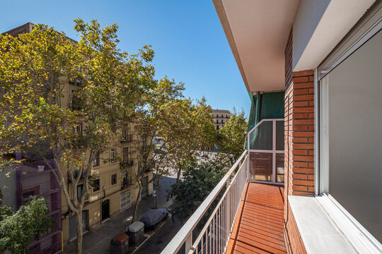 A sunny balcony view overlooking a tree-lined street in an urban area. Bright blue sky and modern buildings are visible