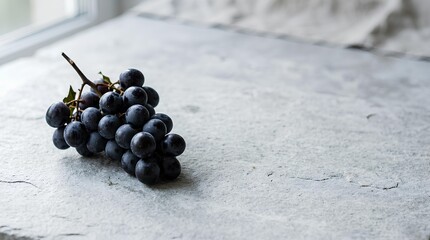 Small cluster of black grapes arranged on the left third of a light gray stone surface, most of the surface left empty as negative space for antioxidant or fruit campaign text, refined and simple.