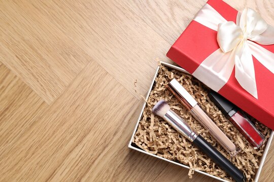 Cardboard gift box with shredded paper filler and makeup products on wooden table, top view. Space for text