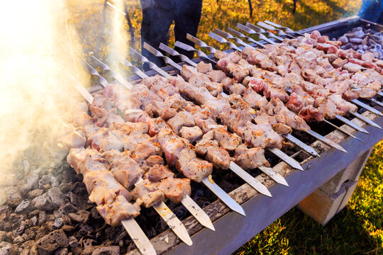 People cook skewers of meat over hot coals at barbecue outside on sunny afternoon.