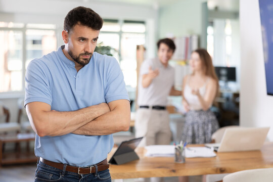Upset young man feeling excluded while other colleagues gossiping behind his back in workplace