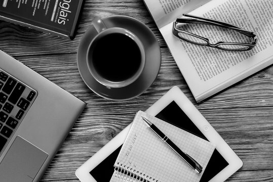 Flat lay of cozy workspace with laptop, red cup of black coffee, open book with glasses and notepad on wooden desk