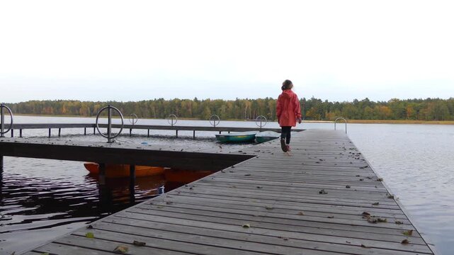 A little girl in a pink raincoat walks along a wooden bridge on the Big Autumn Lake.