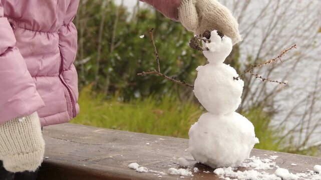 little beautiful girl sculpts a small snowman on a bench near a winter lake