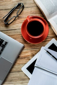 Flat lay of cozy workspace with laptop, red cup of black coffee, open book with glasses and notepad on wooden desk