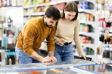 Fototapeta premium Positive young couple choosing cooled food product standing at the fridge in grocery store