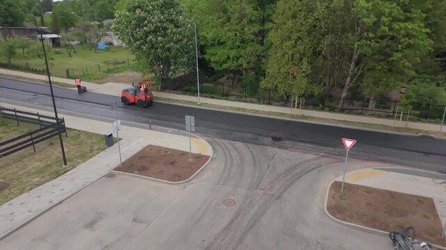 Road construction crew in orange vests operates machinery to lay asphalt on a street in a residential area with greenery