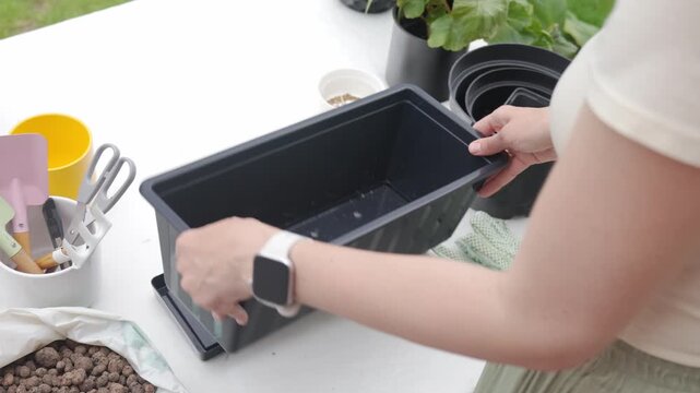 Woman prepares flower pot for container gardening, placing a tray and black planter on a covered table. Concept of planting setup and DIY plant care