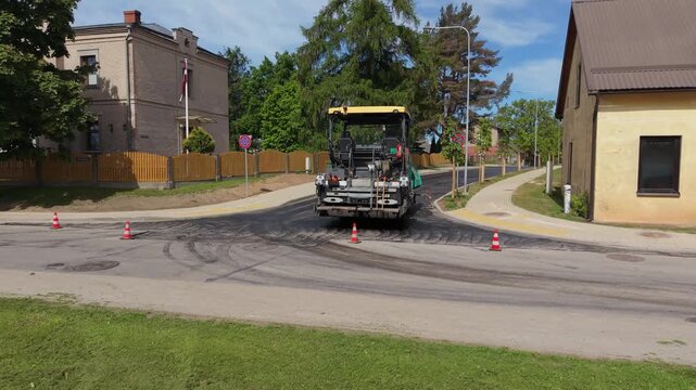 Road construction vehicle laying asphalt on a street corner with traffic cones and residential buildings in the background