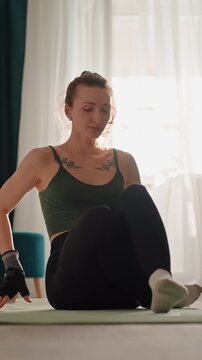 White woman seated spinal twist stretch on mat in sunlit living room wearing green top black leggings socks and gloves gentle mobility routine focusing on posture breath alignment and flexibility